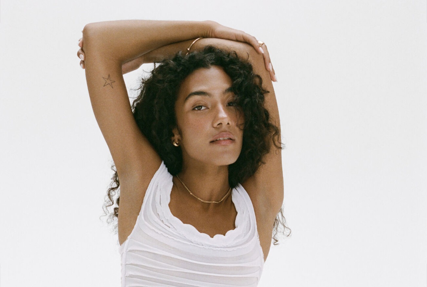 A woman with brown curly hair with a white vest on stood in front of a white background